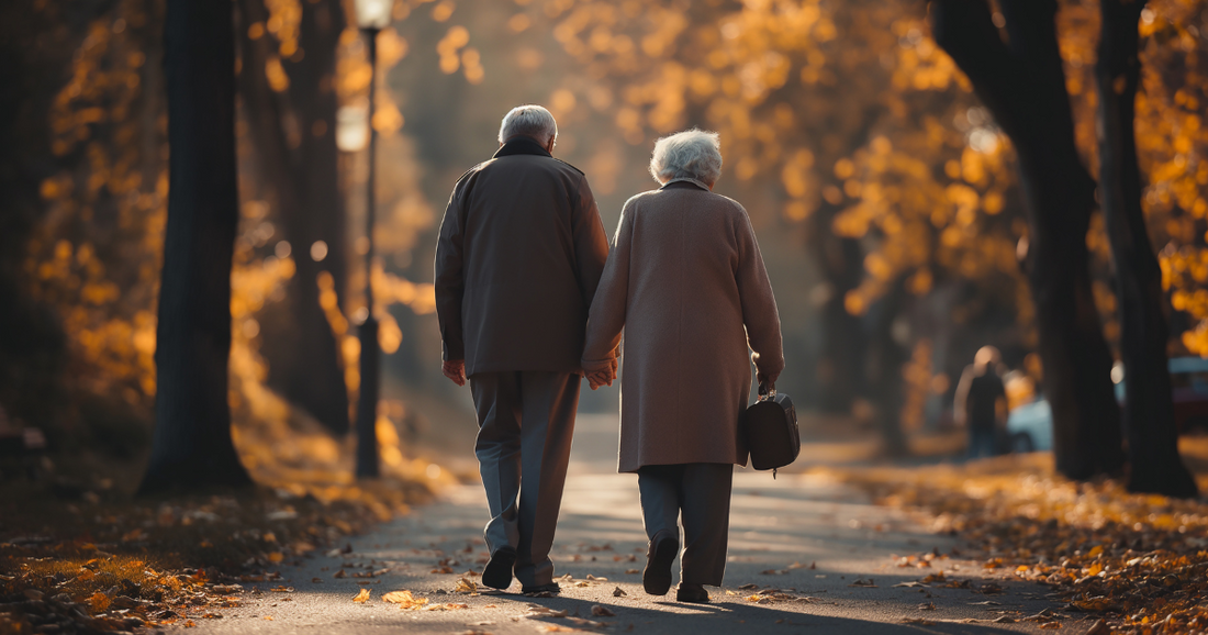 An elderly couple walking hand in hand down a tree-lined path blanketed in golden autumn leaves.