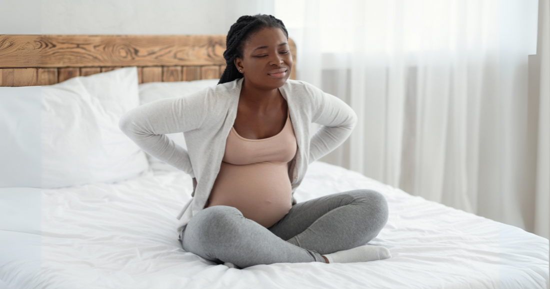 A pregnant woman sits on a white bed holding her lower back with both hands, wearing a pink tank top, gray cardigan, and sweatpants.
