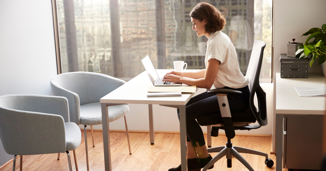 Laptop on a tidy wooden desk with a coffee mug and plant, in a sunlit office.