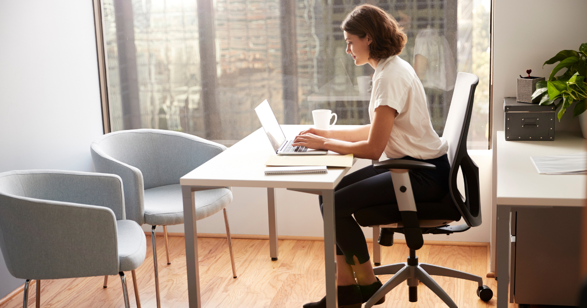 Laptop on a tidy wooden desk with a coffee mug and plant, in a sunlit office.
